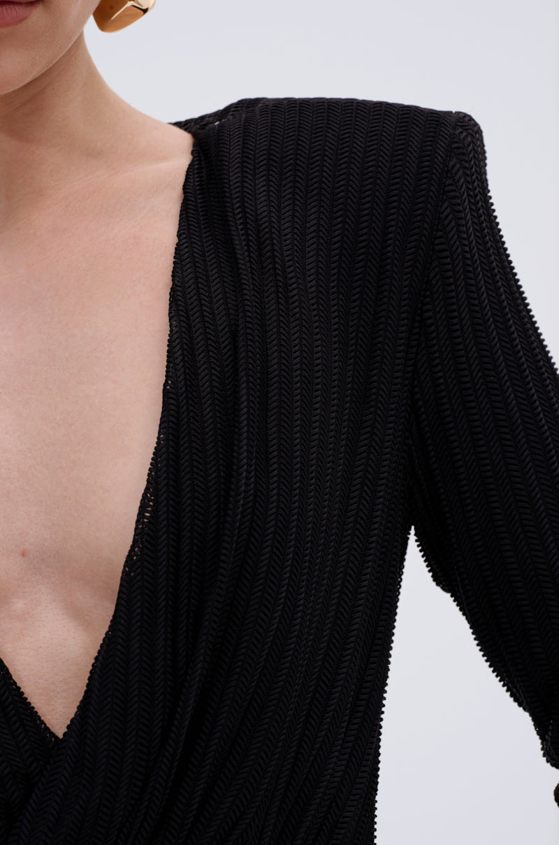A close-up of a person wearing the REBECCA WOVEN MINI DRESS, featuring a textured black fabric and deep V-neckline, paired with a large gold earring against a plain background.