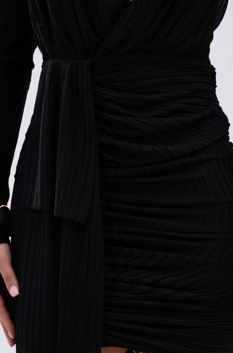 Close-up of a person wearing the REBECCA WOVEN MINI DRESS, a textured black wrap mini dress with a V-neckline and waist tie. Their left arm rests near the hip, showcasing the dress’s horizontal ribbed patterns.