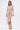 A woman wears the FELT CUTE RUCHED MINI DRESS, a shimmering metallic stretch-knit piece with long sleeves, a deep V-neckline, and an asymmetric draped skirt. She accessorizes with a gold clutch, strappy heels, and large hoop earrings.