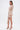 A woman stands against a white background in the FELT CUTE RUCHED MINI DRESS, featuring light beige fabric, a deep V-neckline, and asymmetrical hem. She holds a small clutch and wears strappy heels with leaf details, her long brown hair styled straight.
