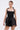 A woman with slicked-back hair wears the LIKE DAYLIGHT SATIN MINI DRESS IN BLACK, a halter-style mini dress with a fitted bodice and flared skirt. She holds a silver clutch and stands against a plain light background.