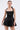 A woman wears the LIKE DAYLIGHT SATIN MINI DRESS IN BLACK, featuring halter straps, a fitted bodice, and a flared skirt, as she holds a geometric metallic clutch against a plain white background.
