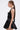 A woman in profile wears the LIKE DAYLIGHT SATIN MINI DRESS IN BLACK, featuring a fitted bodice, flared skirt, and halter straps. She has long brown hair in a low ponytail with hoop earrings against a plain light background.