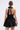 A woman with slicked-back hair, hoop earrings, and a small clutch is shown from behind wearing the LIKE DAYLIGHT SATIN MINI DRESS IN BLACK, featuring halter straps and a large bow at the neck.