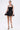 A woman models the LIKE DAYLIGHT SATIN MINI DRESS IN BLACK with halter straps, pairing it with hoop earrings, high-heeled sandals, and a small black clutch against a plain white background.