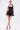 A woman stands against a white background wearing the LIKE DAYLIGHT SATIN MINI DRESS IN BLACK, featuring halter straps and a fitted bodice. She holds a silver clutch, wears bow high-heeled sandals and hoop earrings, with her hair tied back.