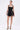 A woman poses against a plain white background wearing the LIKE DAYLIGHT SATIN MINI DRESS IN BLACK with halter straps and a fitted bodice, paired with silver heels and a silver clutch. Her hair is tied back as she looks slightly to the side.