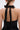 A woman with her hair tied back and hoop earrings models the LIKE DAYLIGHT SATIN MINI DRESS IN BLACK, featuring halter straps, a large satin bow at the neck, and an open back that showcases her shoulders.