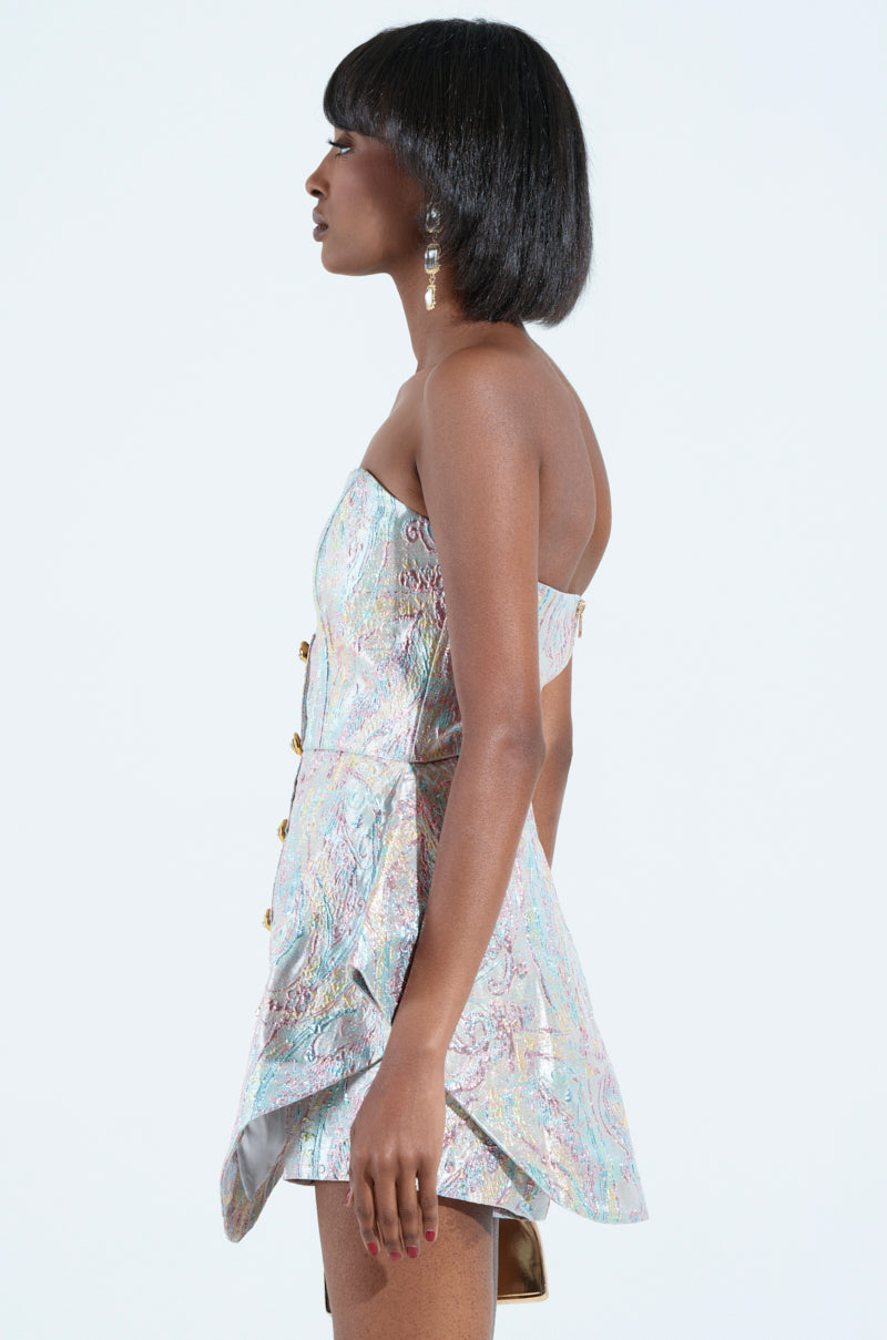 A person with short black hair is shown in profile, wearing the IT GIRL ROMPER IN BROCADE, featuring a strapless sweetheart neckline and iridescent fabric with gold buttons. They are accessorized with dangling earrings against a plain white background.