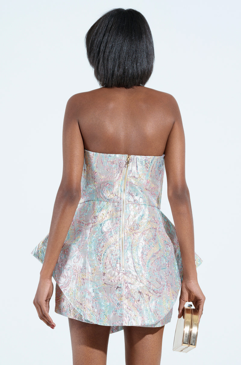 Rear view of a person wearing the "IT GIRL ROMPER IN BROCADE" featuring a strapless sweetheart neckline and pastel colors, with a back zipper. They hold a small, shiny gold purse in their right hand against a plain background.