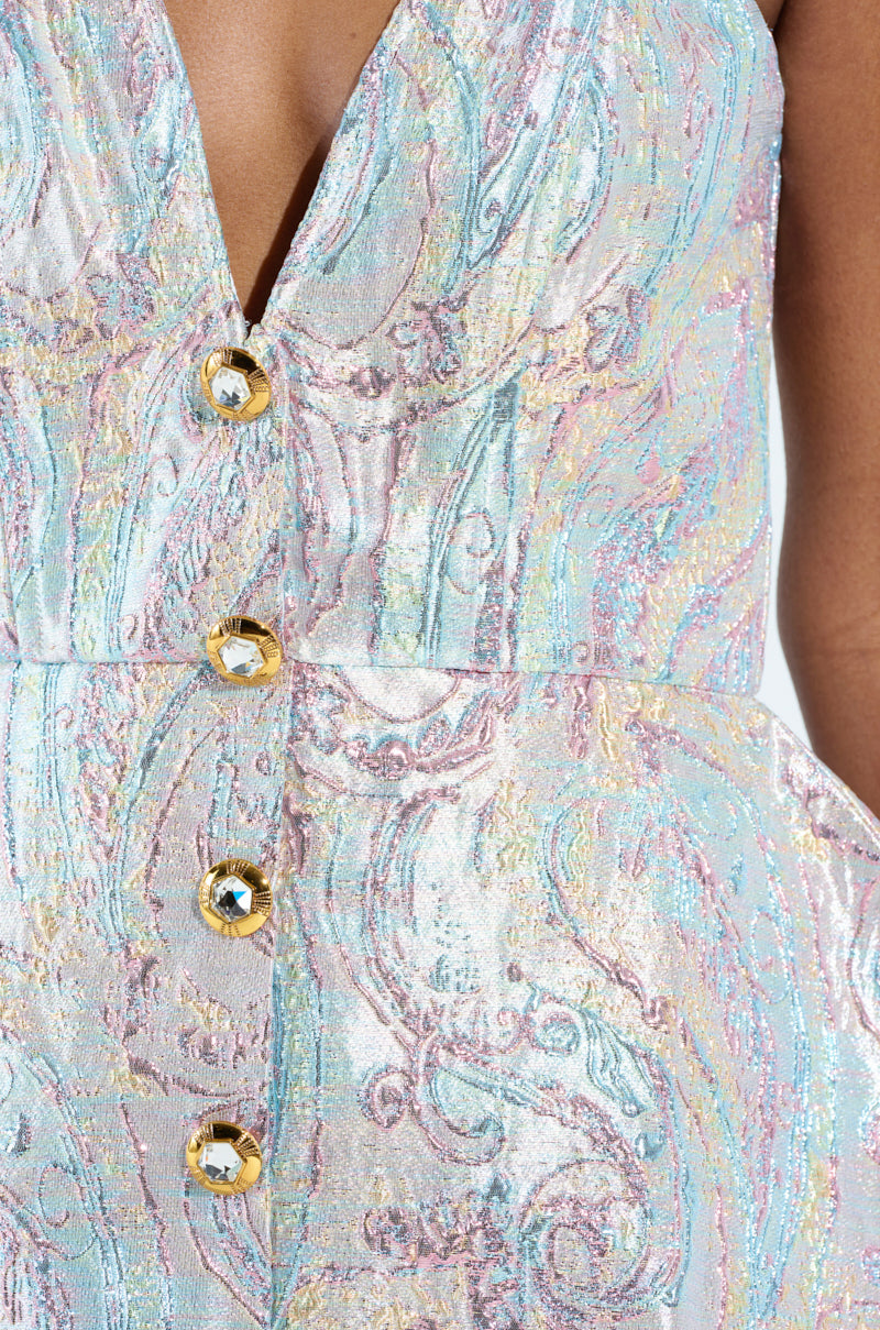 Close-up of a person's torso wearing the IT GIRL ROMPER IN BROCADE with an elegant, strapless sweetheart neckline and ornate patterns. Shiny gold buttons complement the pastel hues of pink, blue, and gold, while the shimmering fabric exudes AKIRA Label's signature luxurious style.