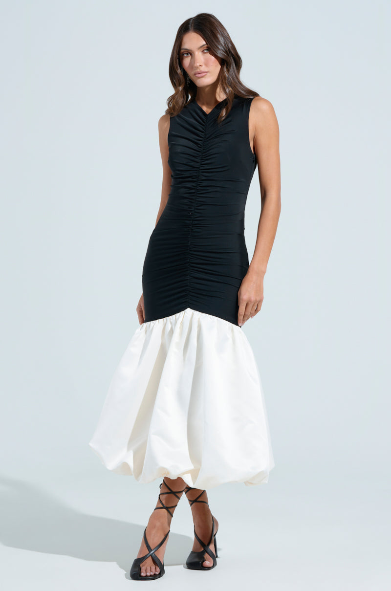 A woman poses in the WITH THE TRENDS BUBBLE MIDI DRESS, featuring a sleeveless black fitted design with a white puffy hem. She stands against a plain background, wearing black strappy heels and her long, wavy hair down.