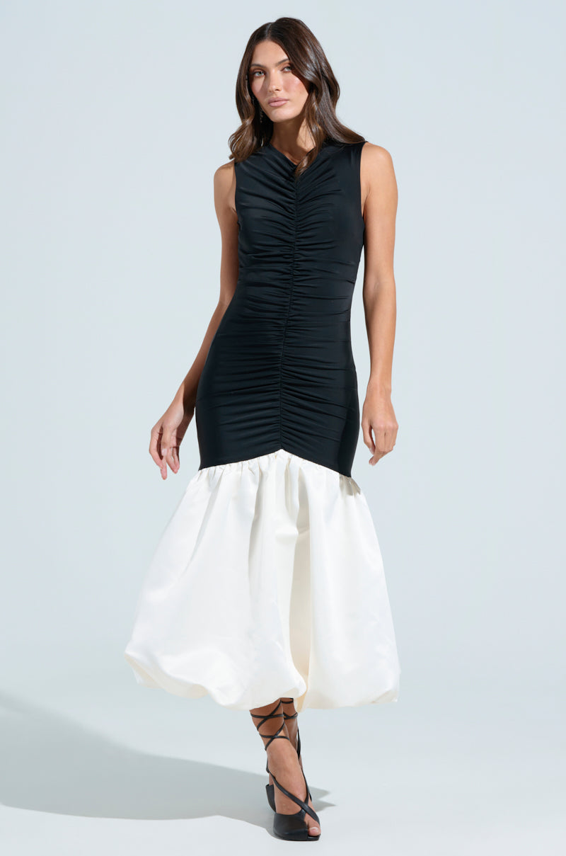 A person wears the WITH THE TRENDS BUBBLE MIDI DRESS, featuring a sleeveless black design with a ruched bodice and voluminous satin skirt. They stand against a plain background, have long brown hair, and wear black strappy heels.