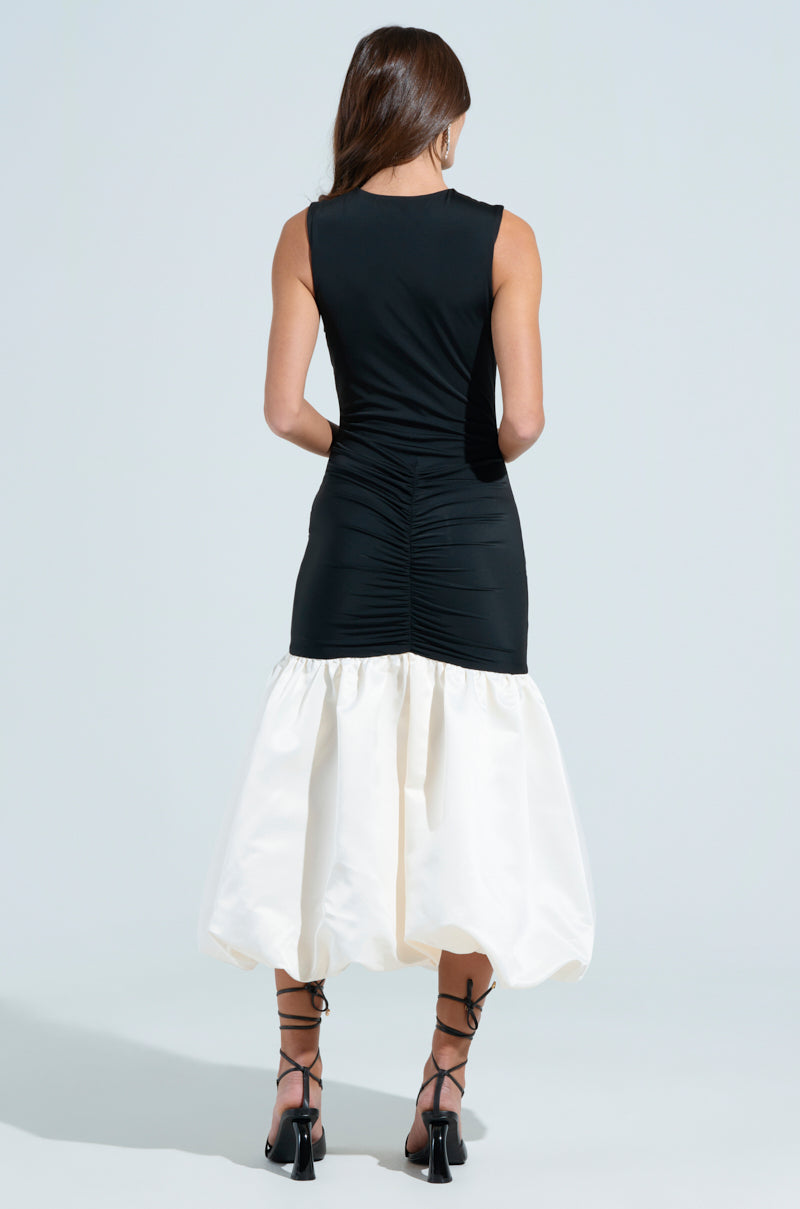 The woman showcases the WITH THE TRENDS BUBBLE MIDI DRESS, a sophisticated sleeveless black dress with a ruched bodice and white satin skirt. Black strappy high-heeled sandals complete her elegant ensemble against a plain, light backdrop.