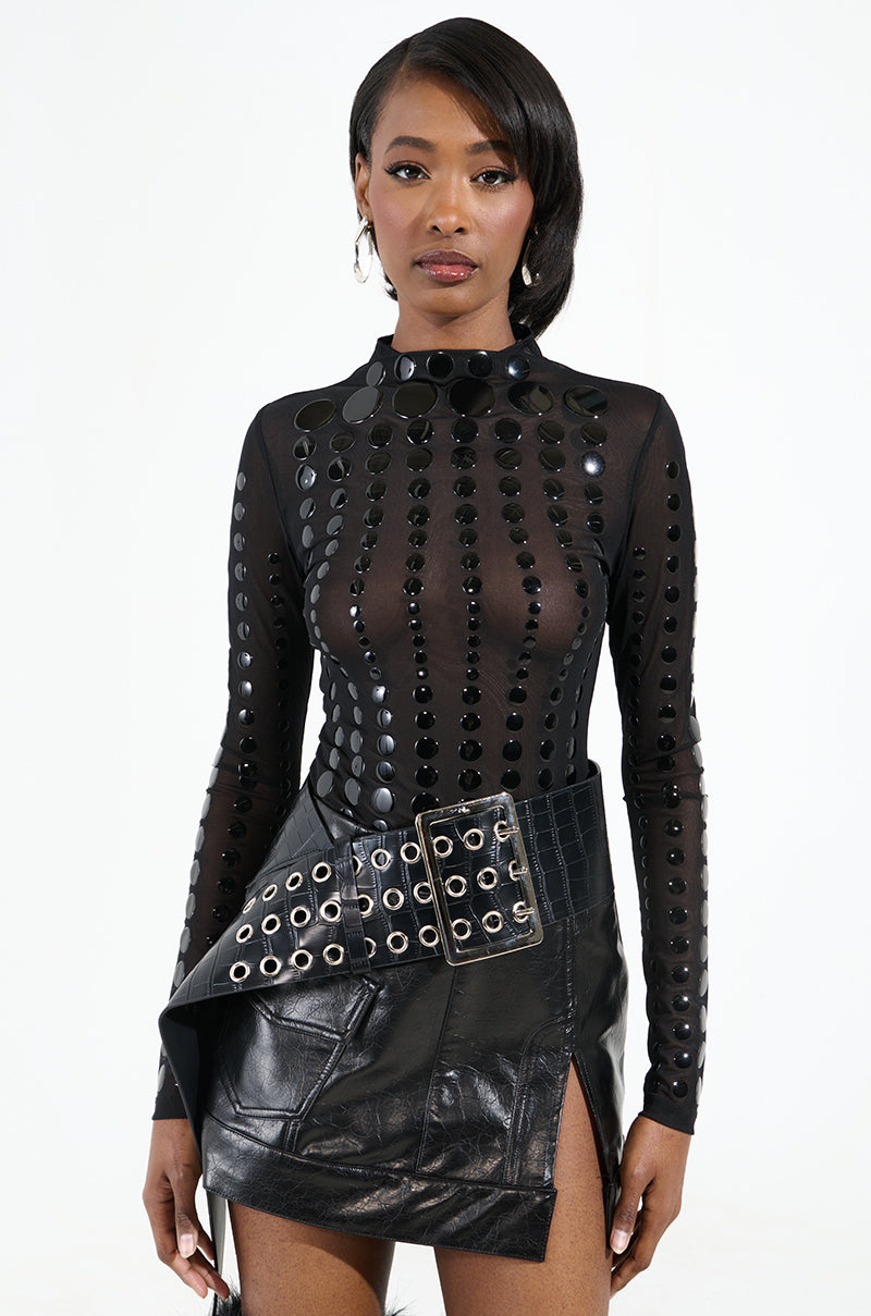 She wears a black, long-sleeved sheer top with circular embellishments and a faux leather skirt featuring grommets paired with the ON TREND OVERSIZED LOW RISE BELT. Her ensemble is finished with long earrings and her hair swept elegantly to one side against a plain white backdrop.