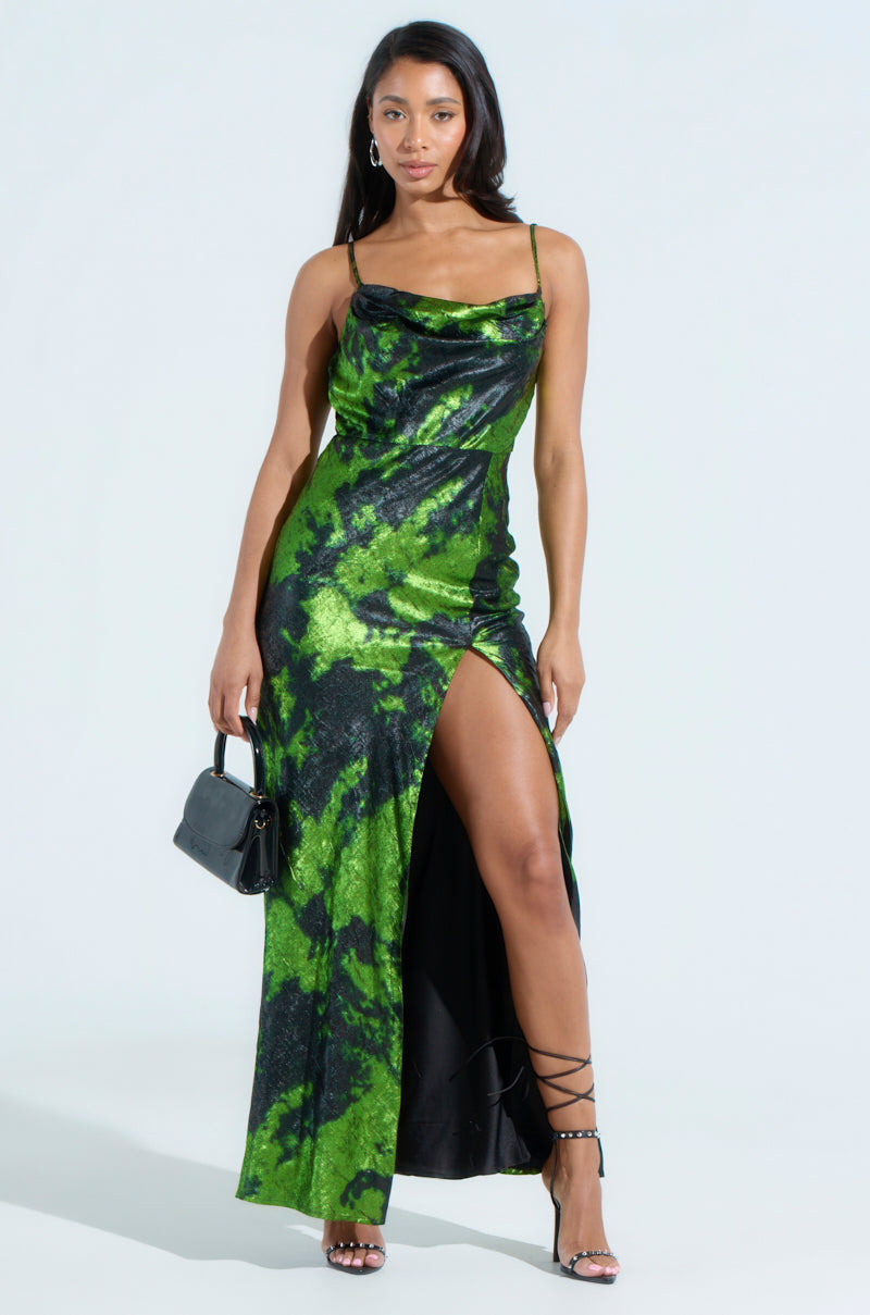 A woman in the LENNON PRINTED METALLIC MAXI DRESS, featuring a sleeveless design and green/black abstract pattern with a front slit, carries a small black handbag and wears strappy heels. Her long hair is loose as she stands against a plain white background.