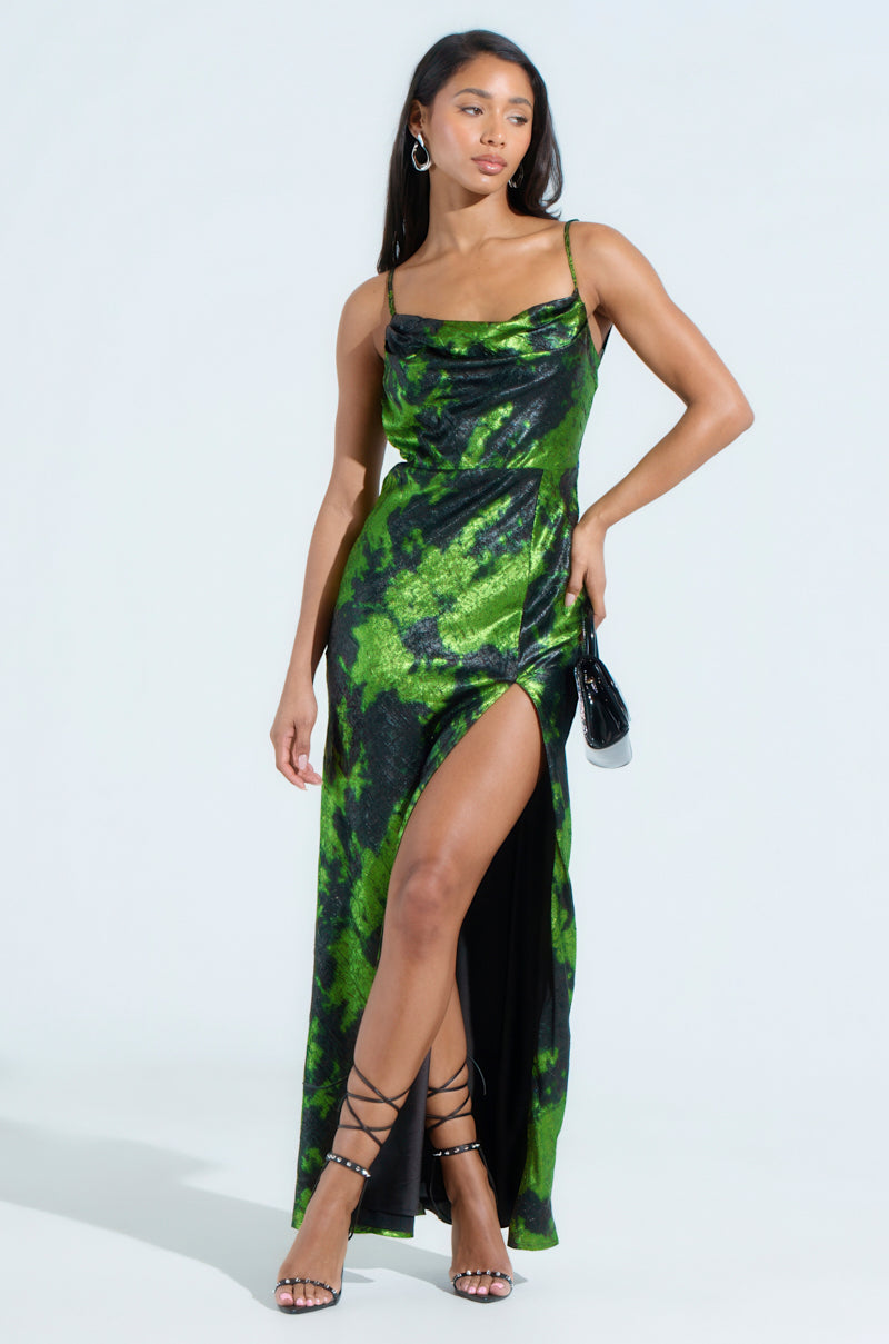 A person poses in the LENNON PRINTED METALLIC MAXI DRESS with a green and black pattern, featuring a plunging front slit. They accessorize with a small black purse and strappy black heels, set against a plain white background.