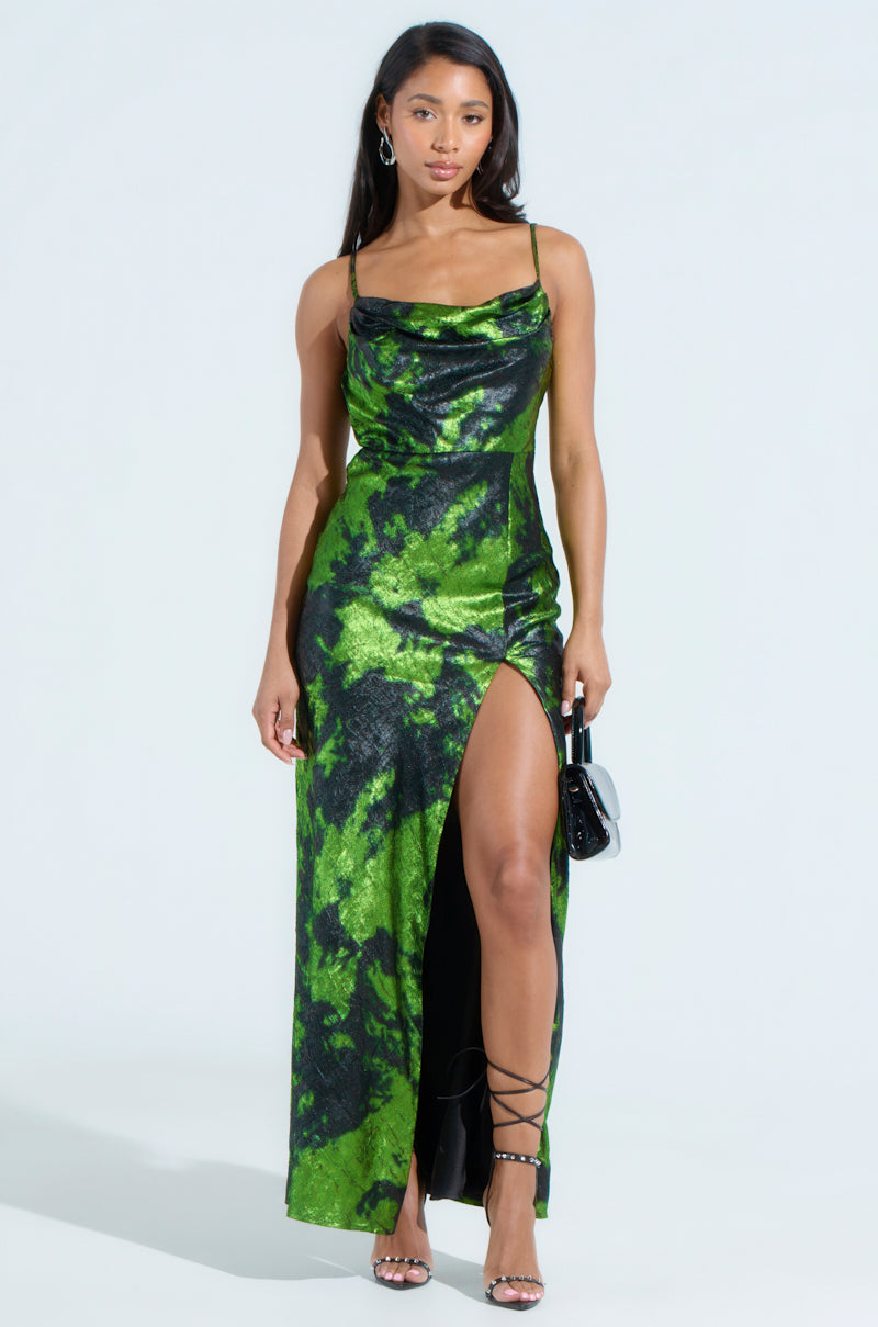 A woman wears the LENNON PRINTED METALLIC MAXI DRESS, featuring a sleeveless design with a green and black marbled pattern and a plunging front slit. She accessorizes with a small black handbag, strappy black heels, long hair down, and glinting hoop earrings against the light gray background.