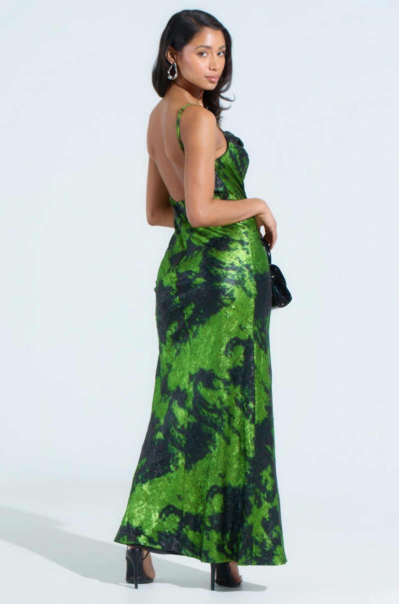 The woman in a LENNON PRINTED METALLIC MAXI DRESS with a low back and plunging front slit turns to the side. She holds a small black purse, wears black heels, and her long hair flows freely. Hoop earrings complete her elegant look.
