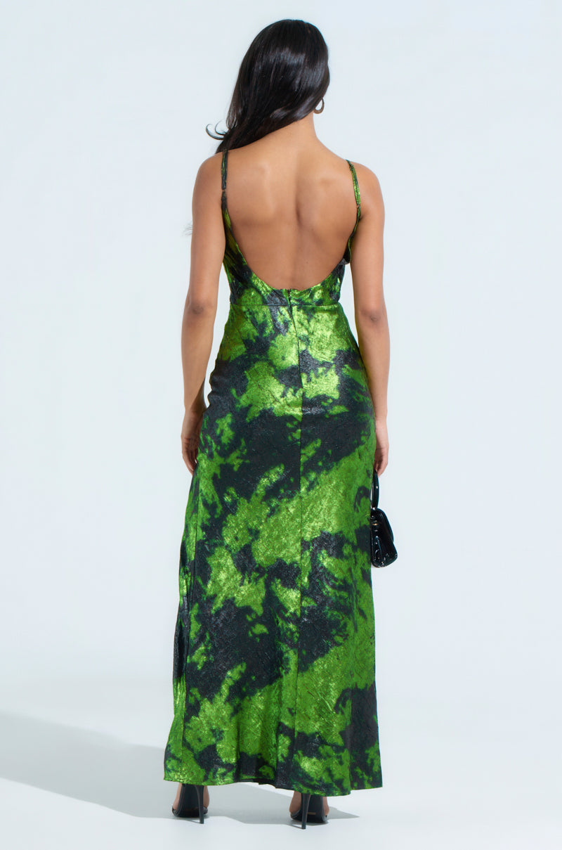 A woman with long hair dazzles in a LENNON PRINTED METALLIC MAXI DRESS featuring a backless design and green and black abstract patterns. The dress flows elegantly as she carries a small black handbag and wears matching shoes, showcased against a plain white background that highlights her stunning style.
