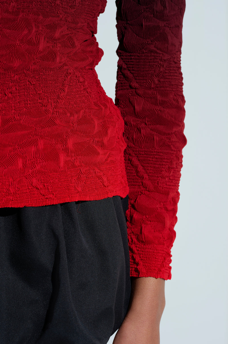 Someone is wearing the BURNING PASSION TOP with its textured red ombre design and loose black pants, their right hand slightly visible by their side against a plain backdrop.