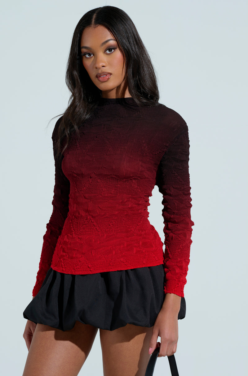 A woman poses against a light background, wearing a BURNING PASSION TOP with a red-to-black ombre design. She pairs it with a figure-hugging black skirt and holds a sleek black handbag, while her long hair flows freely.