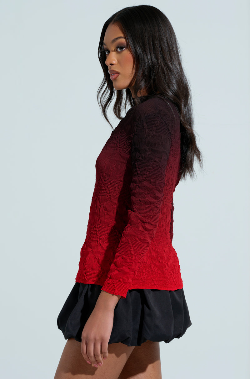A woman with long dark hair wears the BURNING PASSION TOP, showcasing an ombre colorway with black gradient sleeves, paired perfectly with a short black skirt that enhances her curves, as she stands against a light backdrop gazing to the side.
