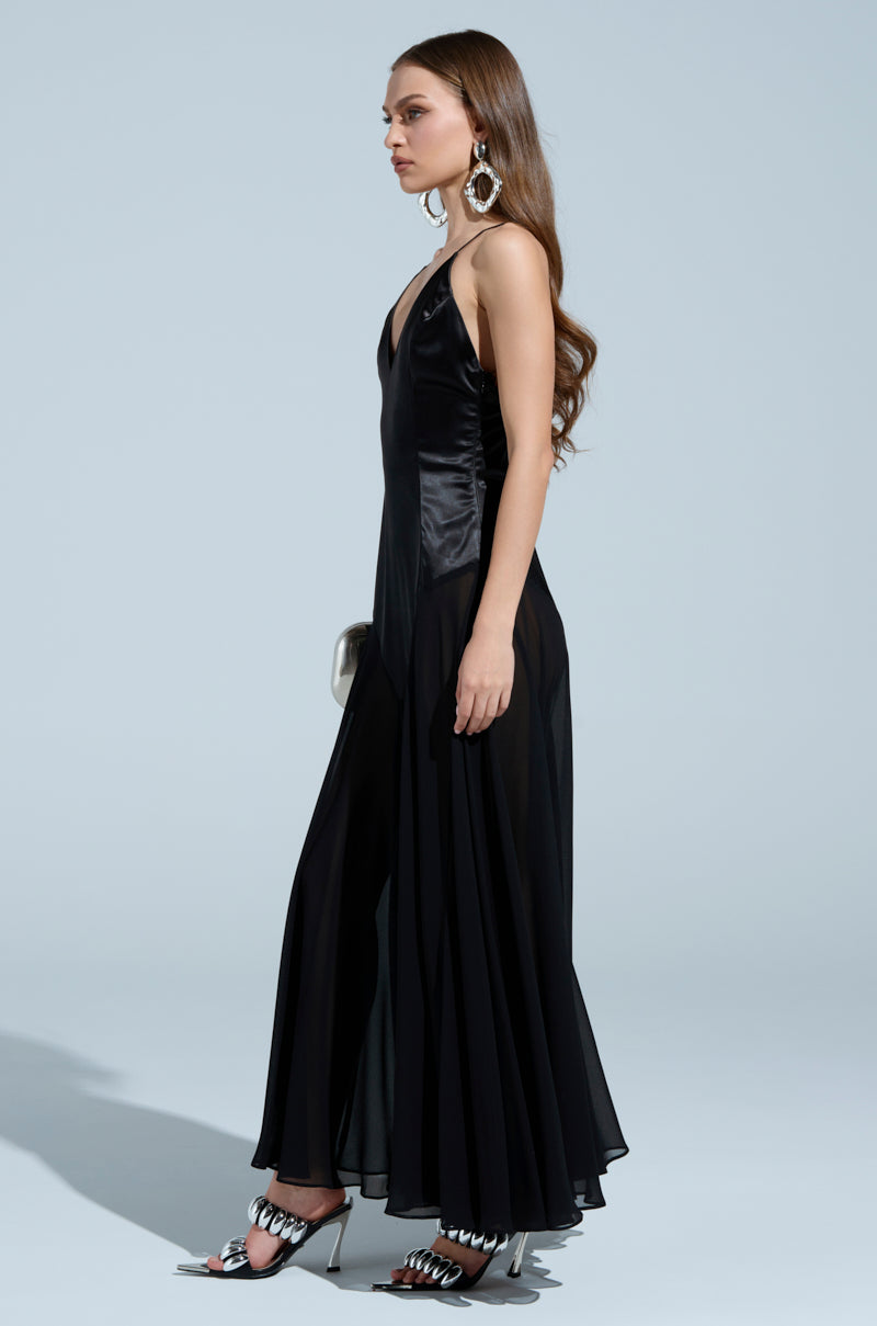 A person with long hair is seen in profile against a plain backdrop, wearing the REINA SATIN MESH MAXI DRESS with adjustable cami straps and a V-neckline. Metallic heels enhance the ensemble, paired with large earrings and a small purse.