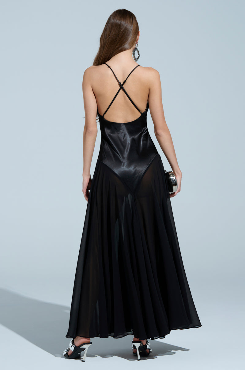 A woman in the REINA SATIN MESH MAXI DRESS, featuring a flowing black design and thin criss-cross straps, stands with her back to the camera. Her long hair complements the V-neckline, while a small clutch adds to her elegance against a plain light gray background.
