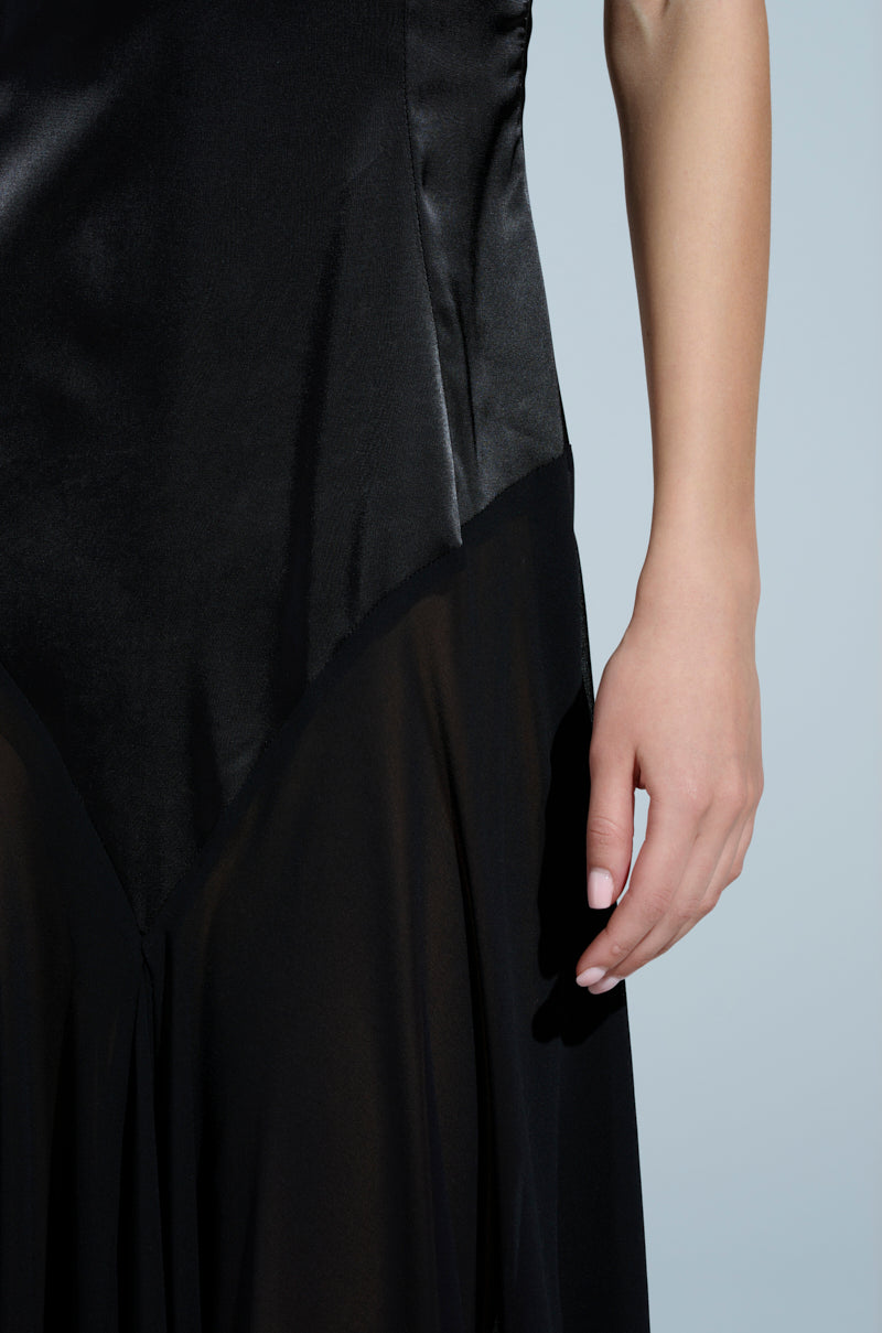 A person is seen in the REINA SATIN MESH MAXI DRESS, featuring adjustable cami straps and a V-neckline. The dress's shimmering satin top transitions to a gauzy lower section. Their hand rests gracefully against a plain, light background.