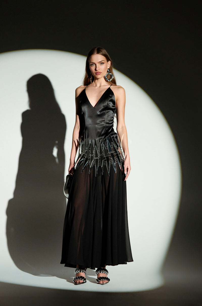 A woman in a REINA SATIN MESH MAXI DRESS with adjustable cami straps and an angular waist design poses against a circular light, her V-neckline highlighting statement earrings while holding a small black purse.