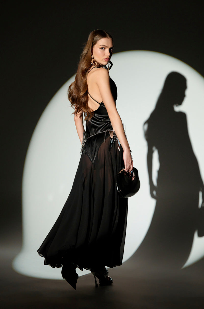 A woman in the REINA SATIN MESH MAXI DRESS with adjustable cami straps poses against a dark background. The V-neckline adds allure while soft light casts a shadow as her long hair cascades down her back. Holding a black handbag, she exudes elegance and moodiness.