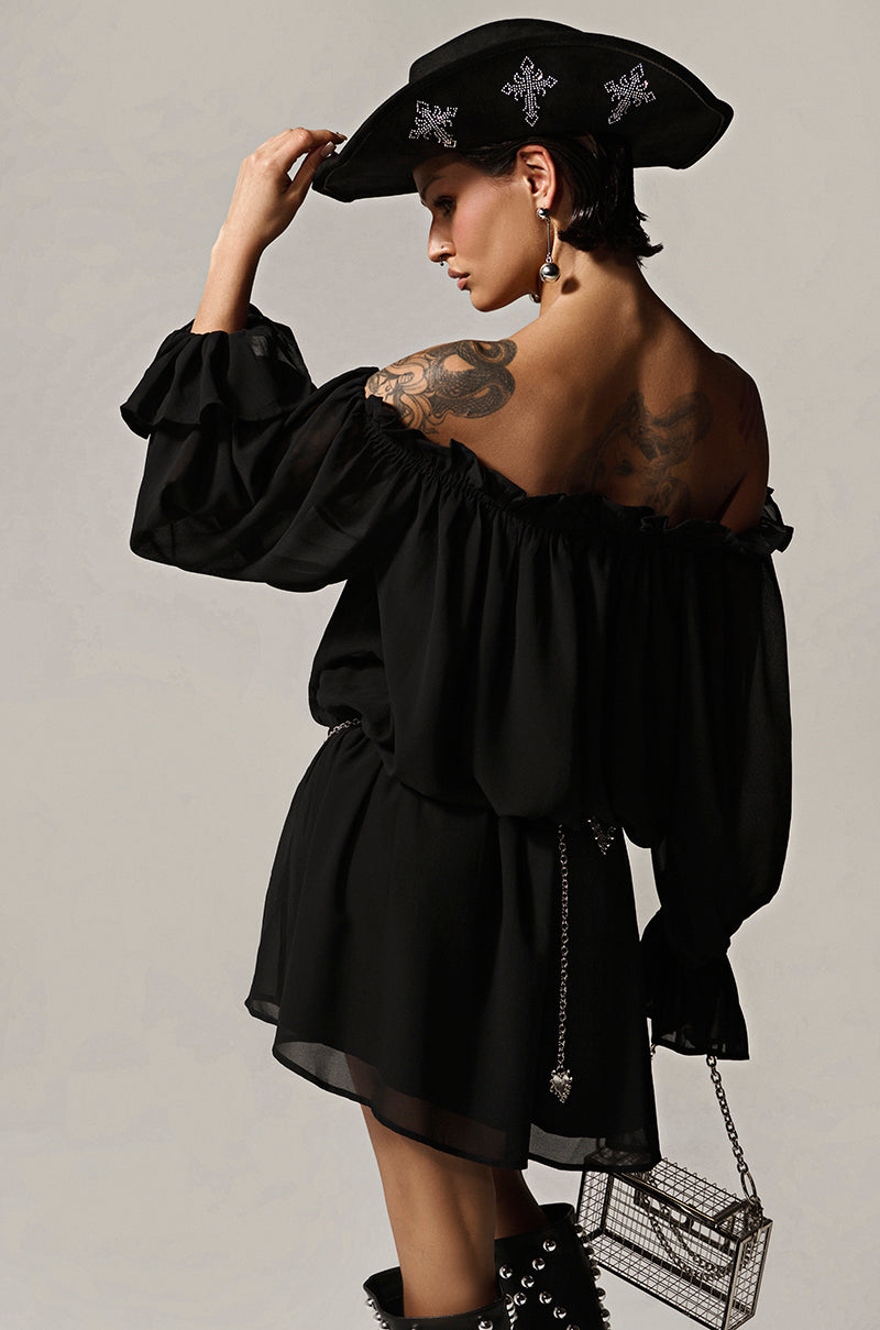 Wearing a CHLOE MESH OFF THE SHOULDER MINI DRESS and a wide-brimmed hat with silver designs, the person faces away, displaying tattoos on their shoulder and arm. Accessories include a chain belt, dangling earrings, and a transparent square handbag.