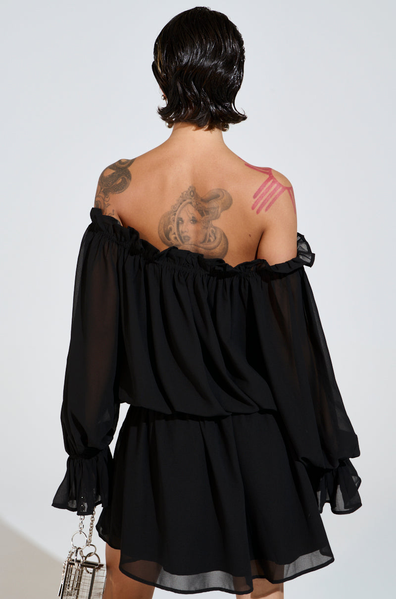 A person with short black hair, seen from behind, wears a chic CHLOE MESH OFF THE SHOULDER MINI DRESS made of chiffon. They have tattoos on their upper back and arm, including a pink abstract design, and hold a small handbag against a plain background.