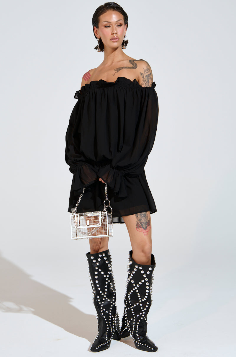 A person stands confidently in a CHLOE MESH OFF THE SHOULDER MINI DRESS, holding a shiny silver handbag and wearing black knee-high boots with large pearls. Tattoos adorn their legs and shoulders against the plain white backdrop.