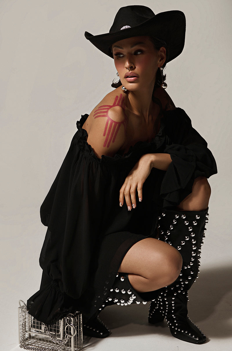 A person in a black Chiffon Chloe Mesh Off The Shoulder Mini Dress poses with one knee bent, sporting a red shoulder design and studded black boots. A small metal cage nearby completes the edgy look.