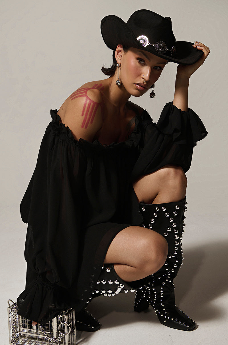 Wearing a CHLOE MESH OFF THE SHOULDER MINI DRESS, silver-studded cowboy boots, and a black hat with silver medallions, a person crouches on a white background. Pink body paint adorns their shoulder, and long earrings enhance the outfit's daring elegance.