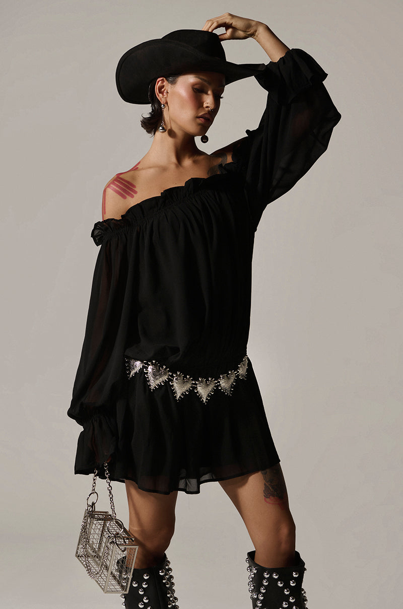 Dressed in a Chloe Mesh Off The Shoulder Mini Dress, they exude confidence paired with a black cowboy hat and studded knee-high boots. A textured handbag and an elasticized waistband belt with silver accents complete the look, as their left arm gently touches the hat's rim.