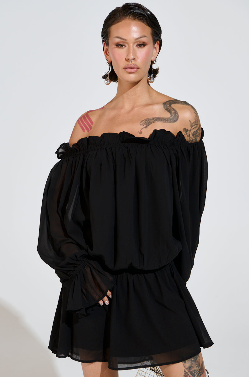 Wearing a CHLOE MESH OFF THE SHOULDER MINI DRESS, a person poses against a simple backdrop. The black chiffon drapes elegantly on their frame, complementing their short dark hair and arm tattoos, exuding confidence with one hand resting gently on the dress.