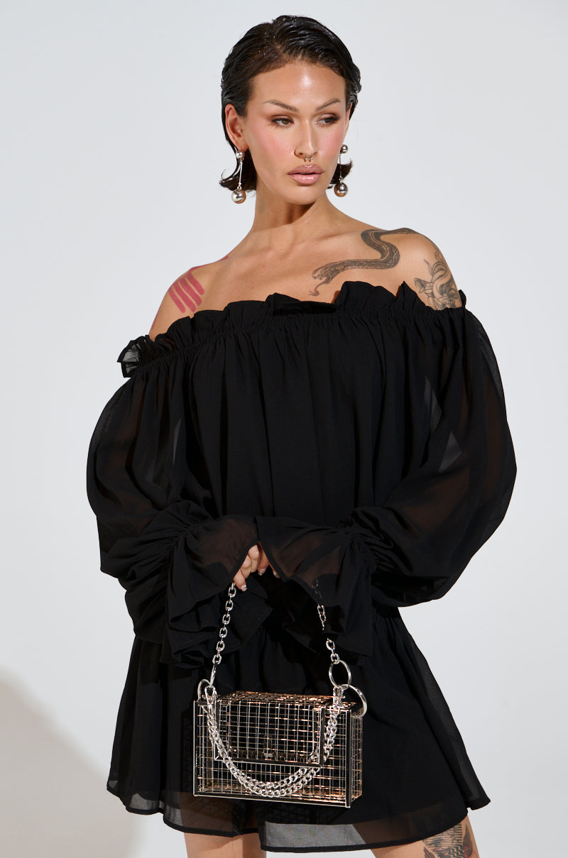 In a black Chloe Mesh Off The Shoulder Mini Dress with long sleeves and an elastic waistband, a tattooed person stands poised, hair styled back. They accessorize with a silver chain bag and statement earrings.