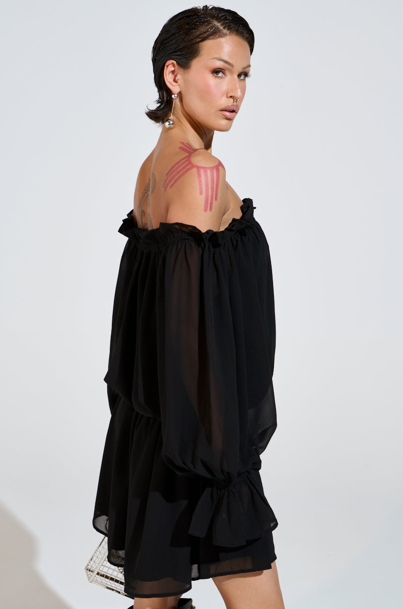 With short hair, a person poses backwards in the CHLOE MESH OFF THE SHOULDER MINI DRESS complete with a ruffled hemline. Holding a small purse, they showcase pink paint on their shoulder and wear dangling earrings.