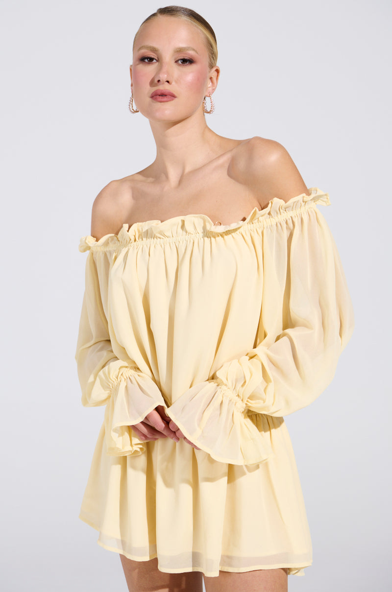 A person with blonde hair in an updo wears the ON YOUR MIND CHIFFON MINI DRESS, featuring a light yellow off-the-shoulder design with ruffled edges. They accessorize with hoop earrings, posing against a plain backdrop for an elegant and timeless look.
