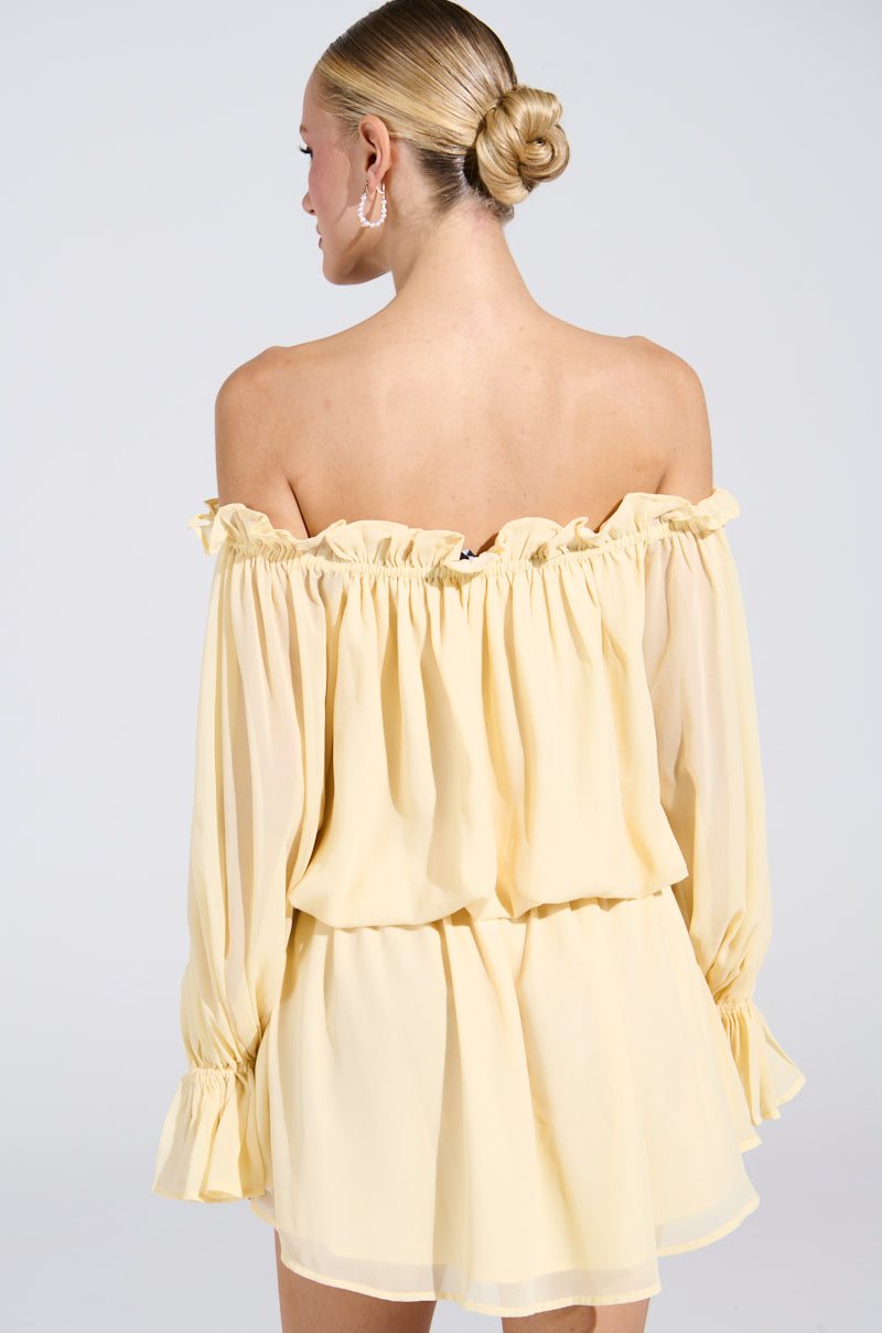 A woman with blonde hair in a bun dons the ON YOUR MIND CHIFFON MINI DRESS, featuring an off-the-shoulder, light yellow design with ruffled edges and long sleeves. She faces away, highlighting the dress's back against a plain white background.