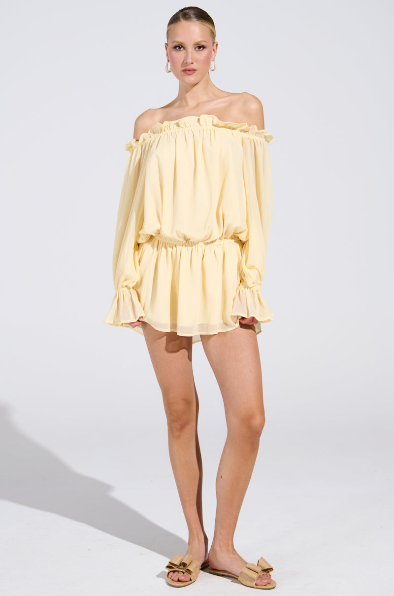 The person is in front of a plain backdrop, donning an ON YOUR MIND Chiffon Mini Dress in light yellow pastel with an off-the-shoulder design, featuring a ruffled neckline and hem. They elegantly pair it with tan slip-on sandals and hoop earrings.