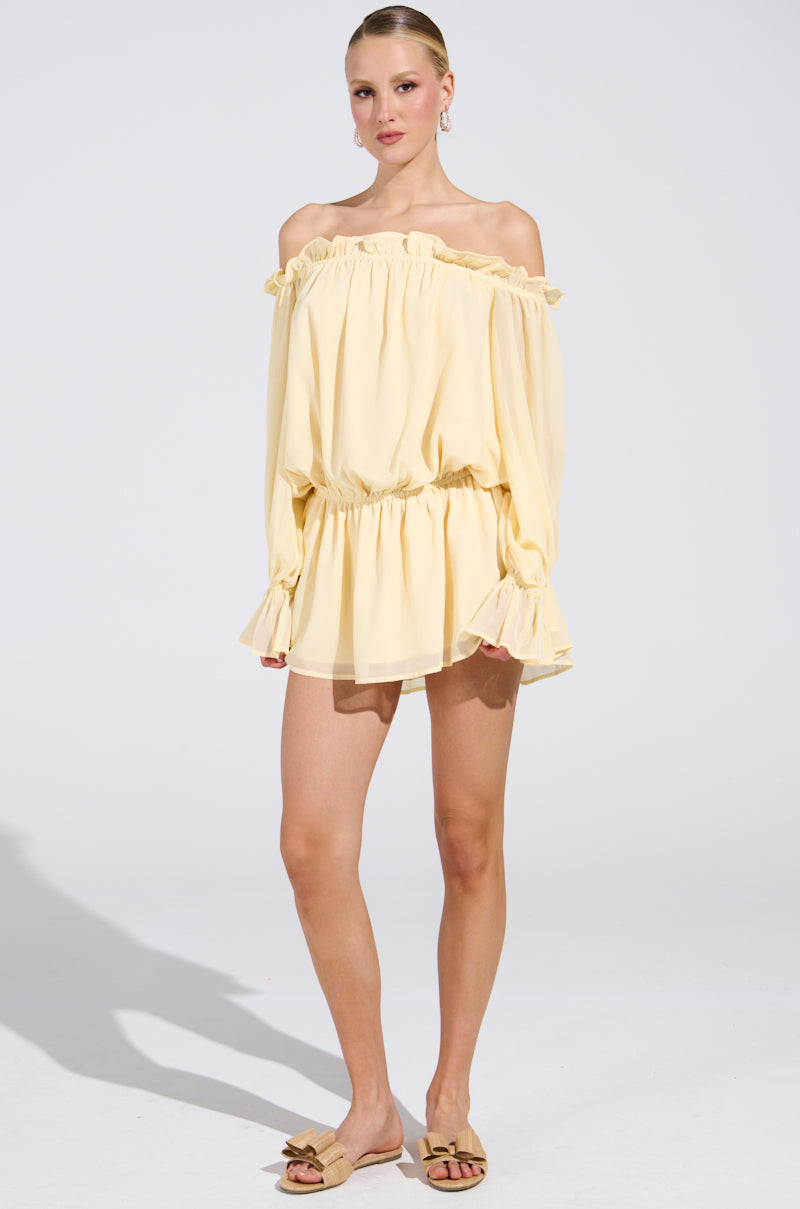 A person in the "ON YOUR MIND" light yellow chiffon mini dress, featuring an off-the-shoulder style with long sleeves, ruffled neckline, and elastic waist, stands against a white background. Their hair is pulled back as they wear tan sandals.