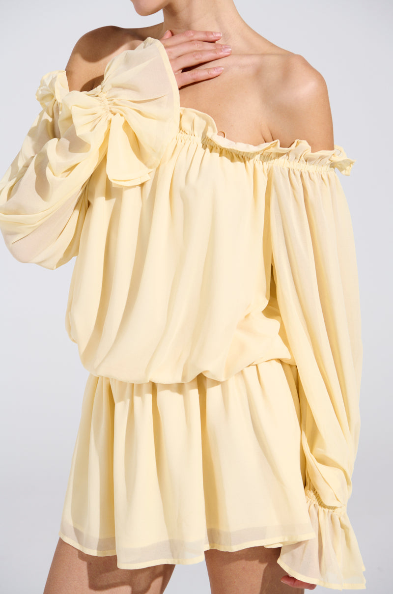 A person models the ON YOUR MIND CHIFFON MINI DRESS, an off-the-shoulder yellow mini dress with voluminous long sleeves, ruffled neckline, and gathered waist, posing with one hand near their chin against a neutral background.