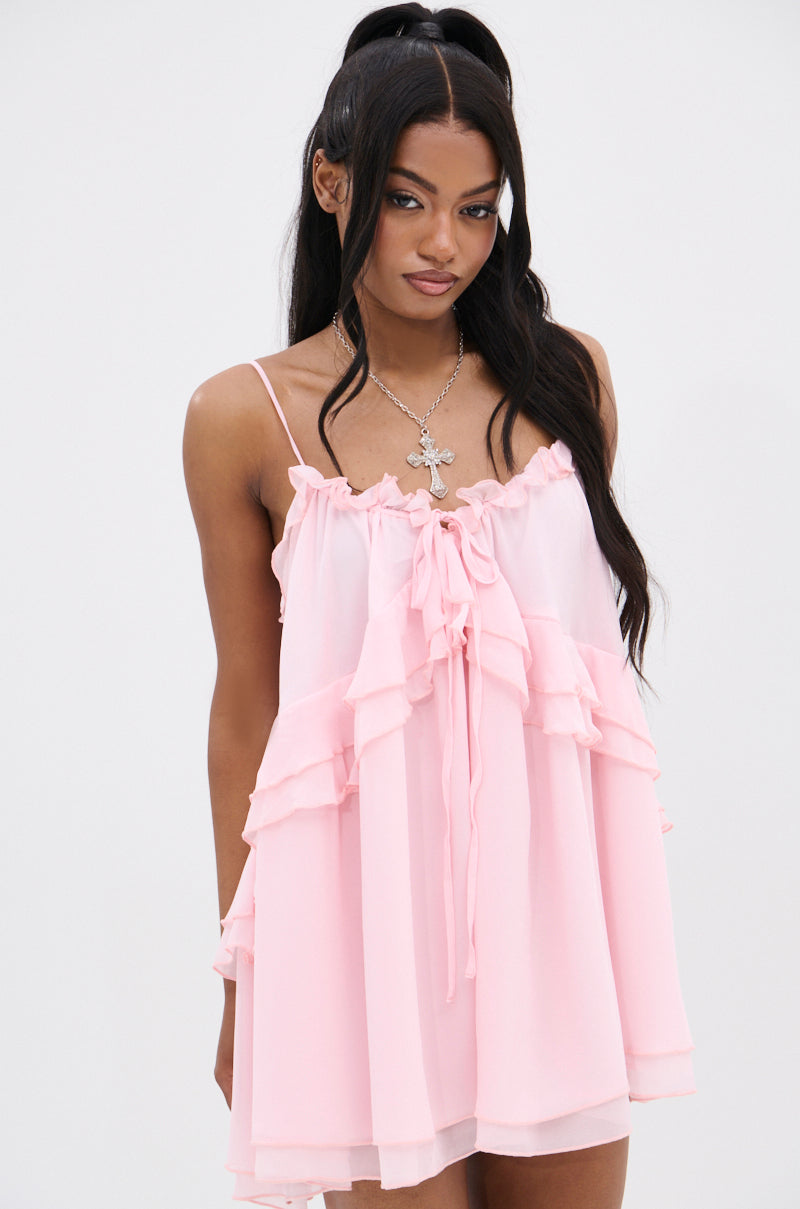 Against a white backdrop, a person with long dark hair styled in two ponytails poses wearing the VANJA Chiffon Mini Dress, featuring sleeveless design, ruffled pink chiffon fabric, and adjustable tank straps. A cross necklace completes the effortlessly chic look.