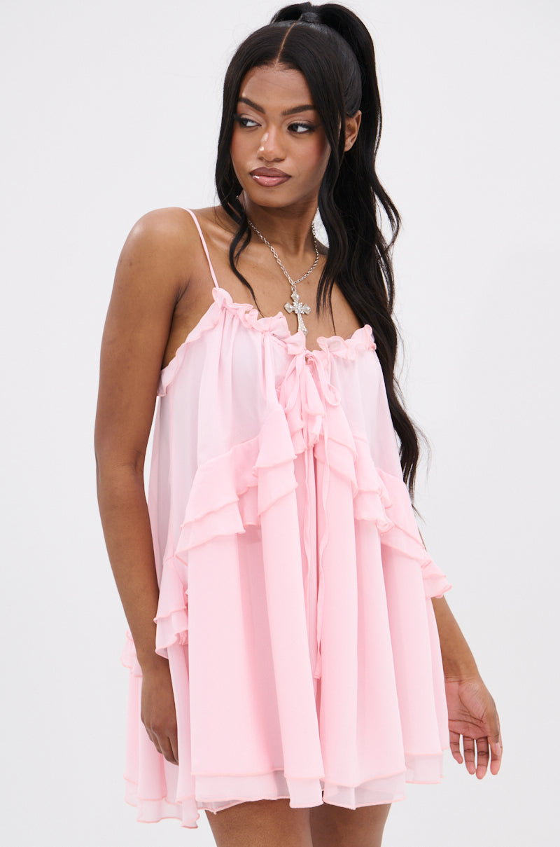 A woman in the VANJA CHIFFON MINI DRESS, featuring a pink hue with a ruffle-trimmed V-neckline and adjustable tank straps, gazes sideways. Her long dark hair is styled in a high ponytail, perfectly showcasing her cross necklace against the plain white background.