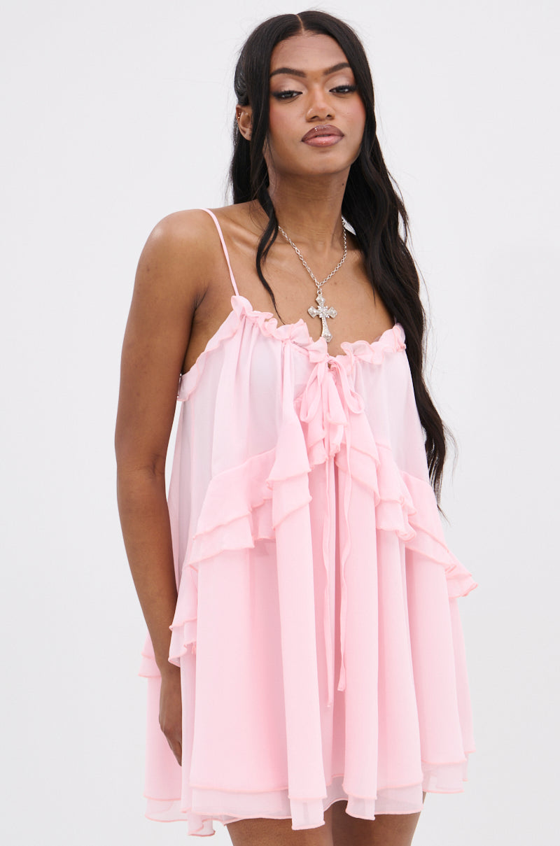 In a VANJA CHIFFON MINI DRESS, a woman adorned in pink chiffon with adjustable tank straps and a front bow pairs her ruffle-trimmed V-neckline with a cross pendant necklace. Her long dark hair elegantly contrasts against the plain white background.
