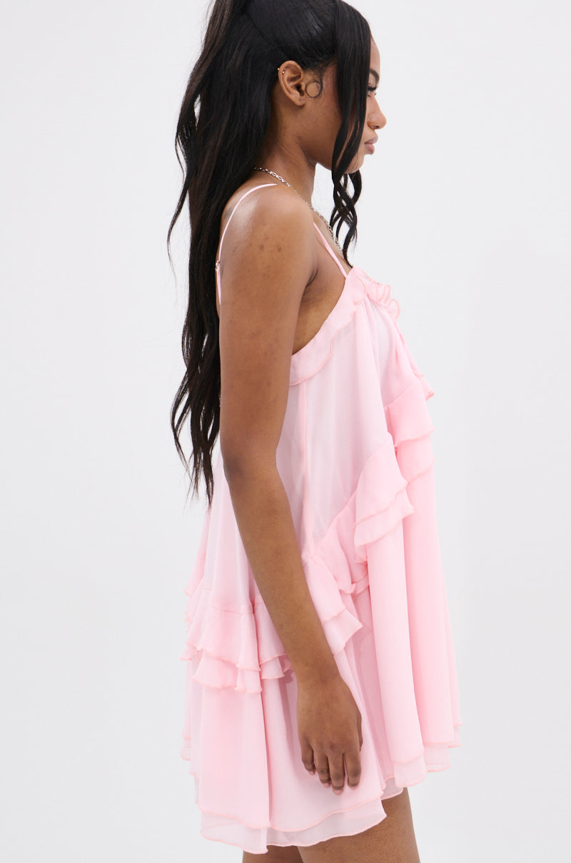 A woman with long, dark hair poses sideways in the VANJA CHIFFON MINI DRESS, showcasing its ruffled, flowing layers and adjustable tank straps. The simple white backdrop accentuates the dress's elegant charm.